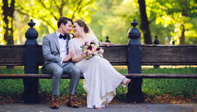 groom and bride with wedding bouquet sitting on a park bench